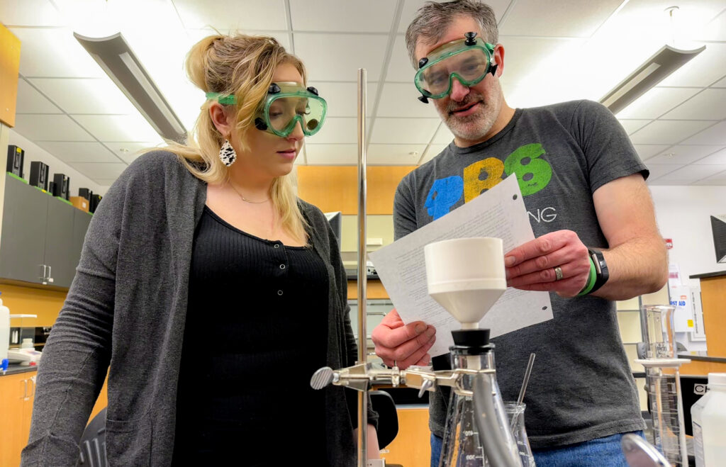 Photo of chemistry instructor Dan Casmier looking at a sheet of paper with a student. Both are wearing safety goggles.